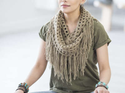 A Caucasian woman is indoors in a fitness studio. She is wearing casual exercise clothing and a scarf. She is sitting cross-legged on a yoga mat and meditating with her eyes closed.